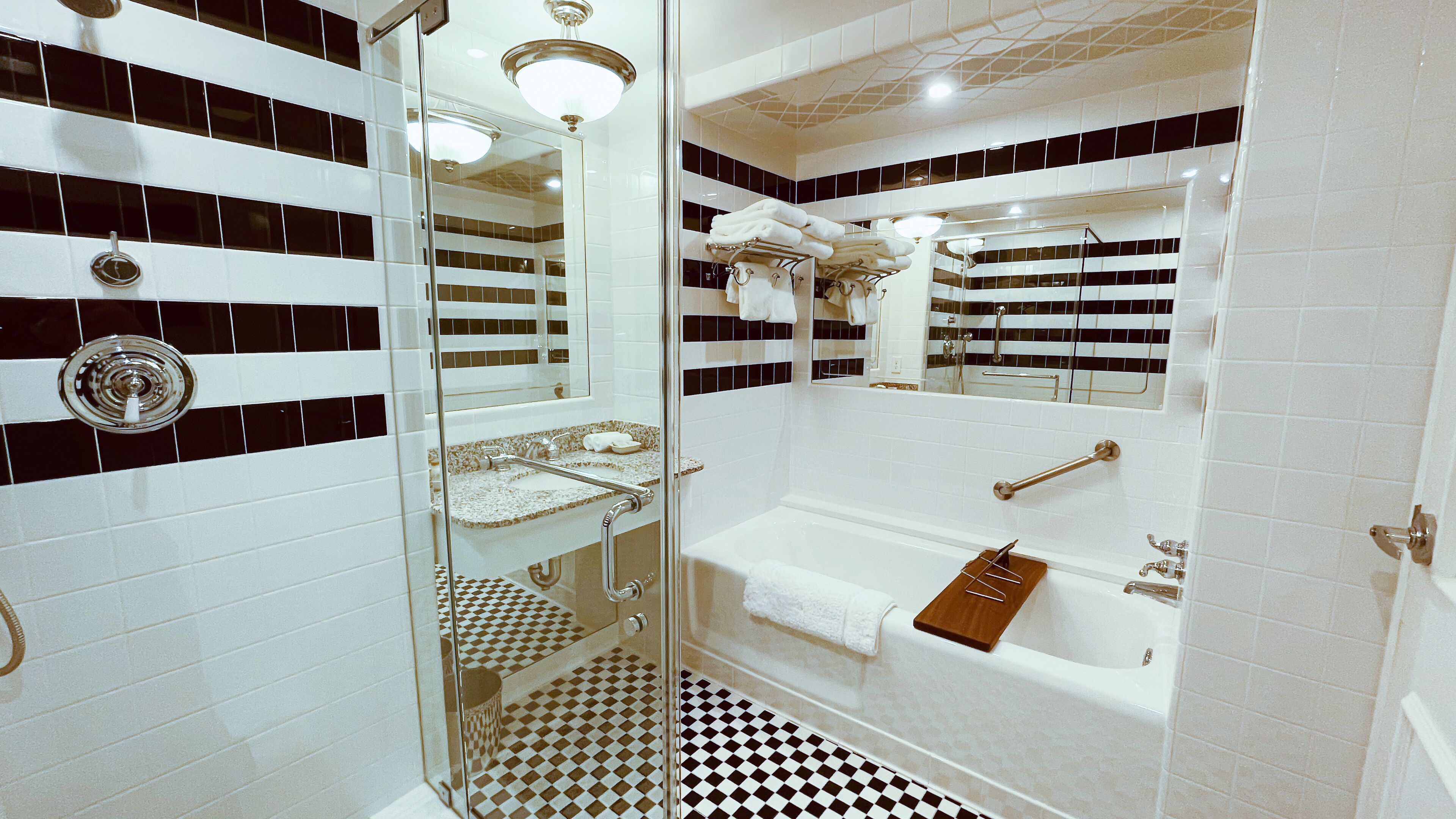Un baño con piso a cuadros, ducha con cerramiento de vidrio, lavamanos con barra para toallas y bañera con bandeja de madera.