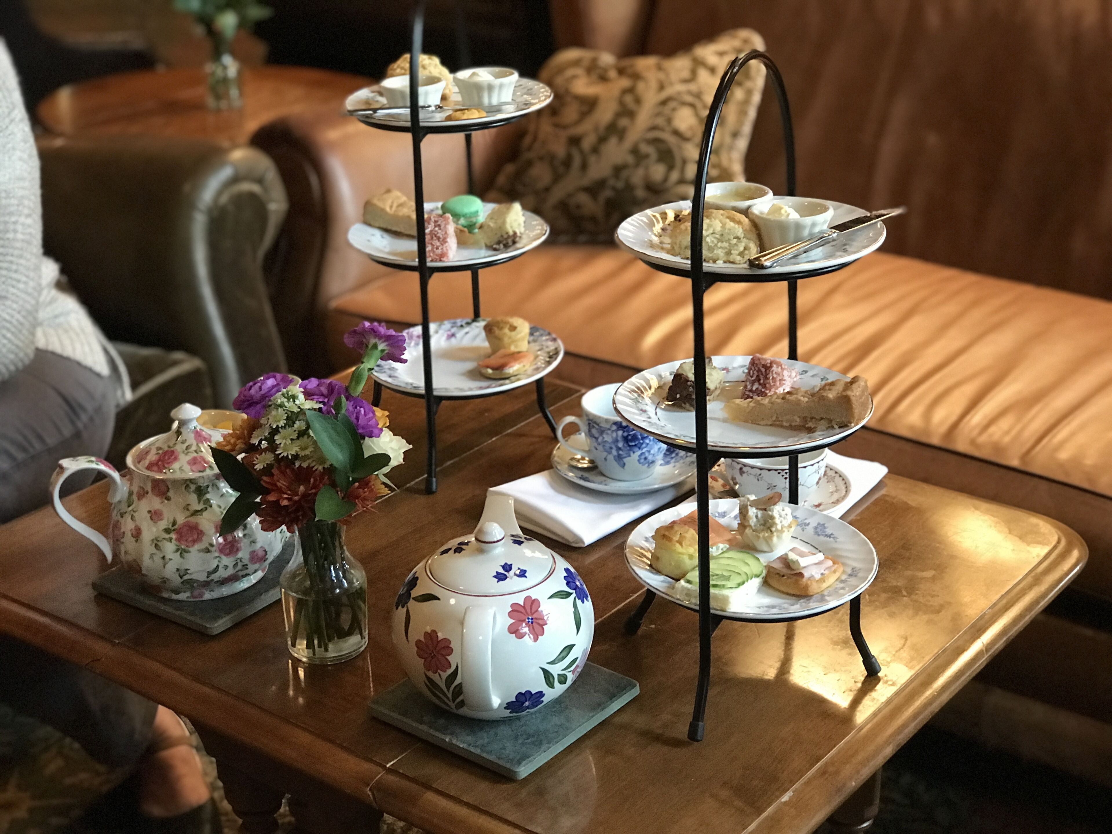 A three-tiered tea service set on a table with floral-patterned teapots and cups, accompanied by a vase of flowers.