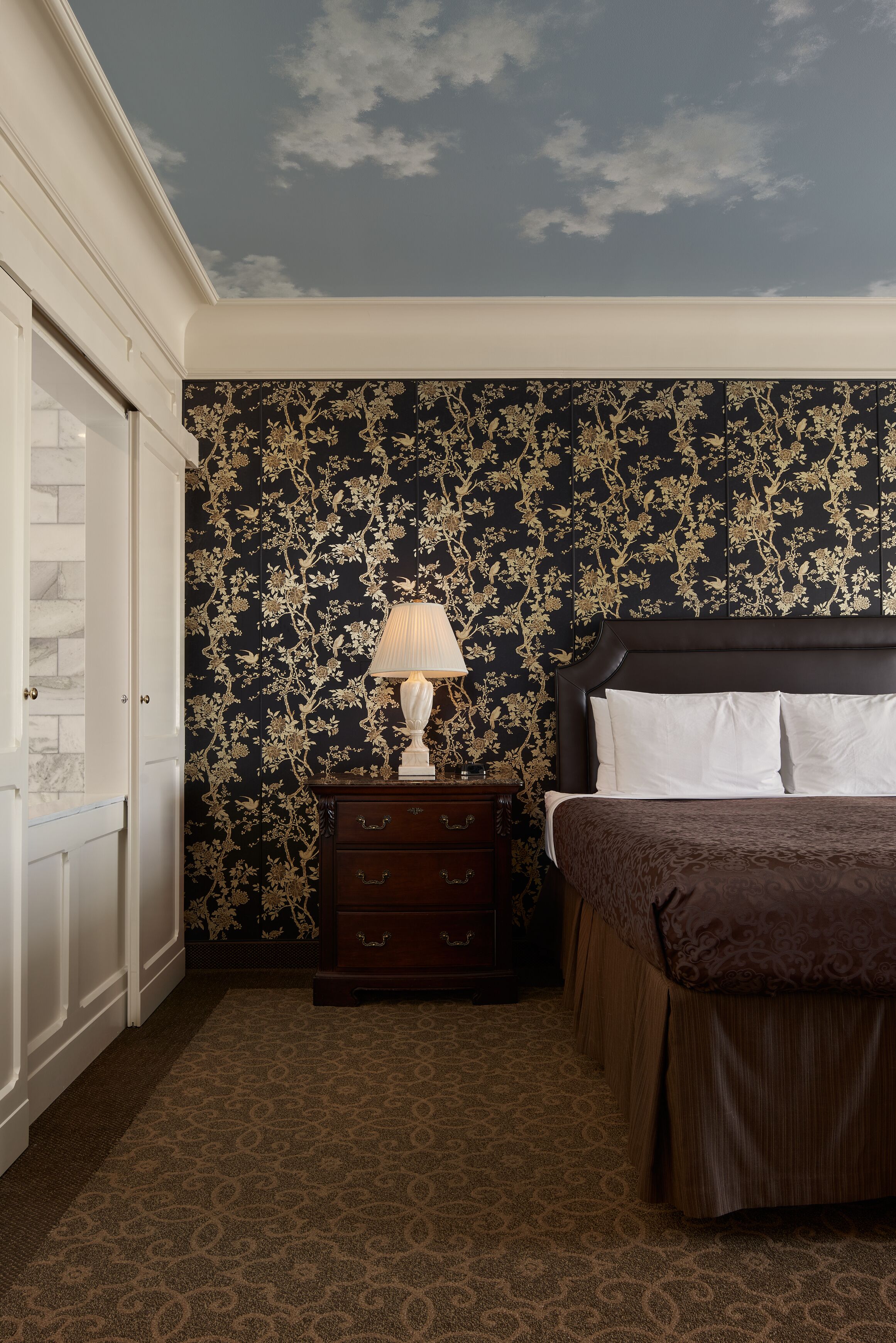 A hotel room with a patterned wallpaper, a bed with white linens, a nightstand with a lamp, and a dark wooden dresser.