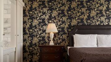 A hotel room with a patterned wallpaper, a bed with white linens, a nightstand with a lamp, and a dark wooden dresser.