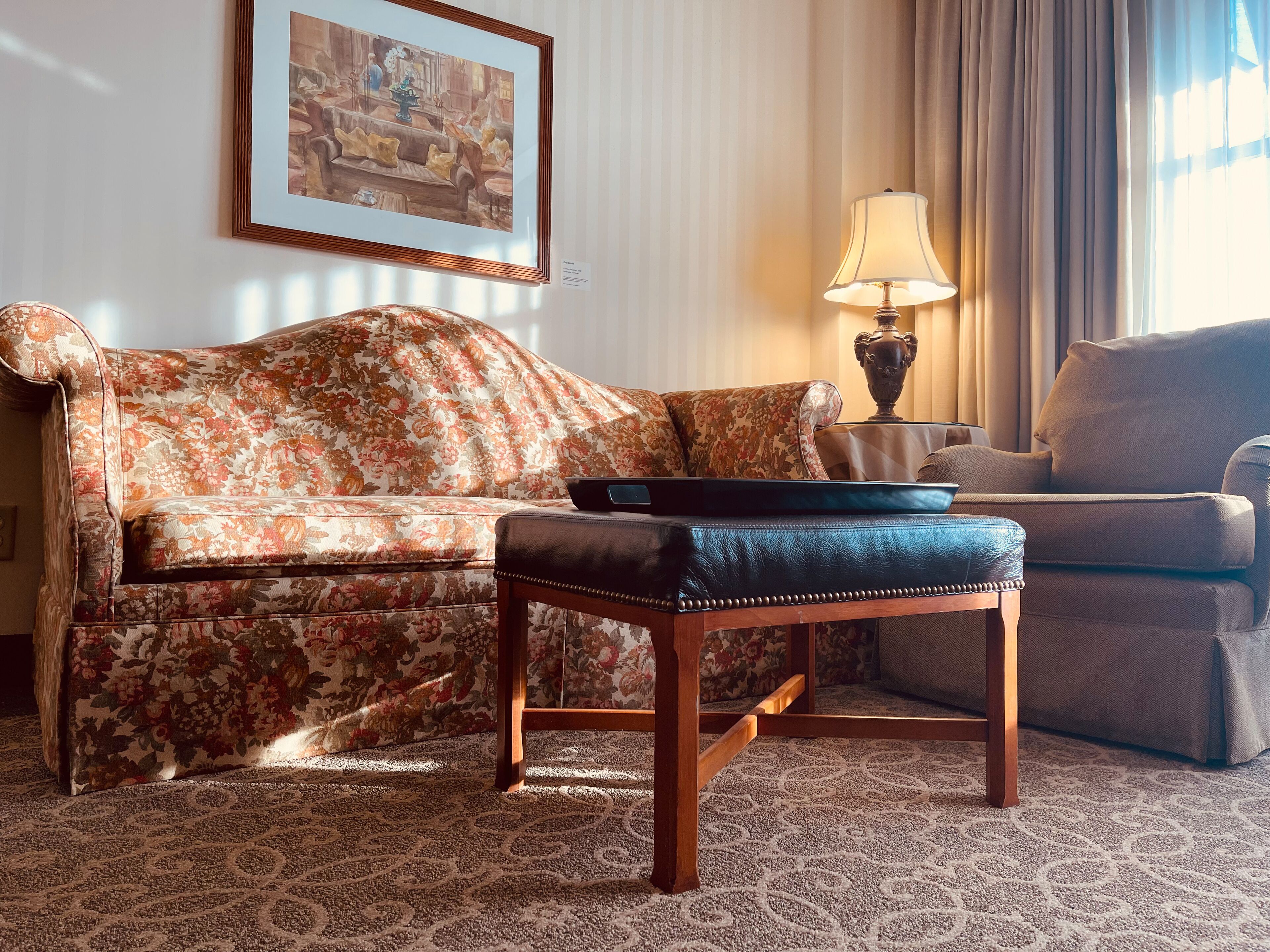 A living room with a floral sofa, a matching ottoman, a side table with a lamp, and a framed painting on the wall.