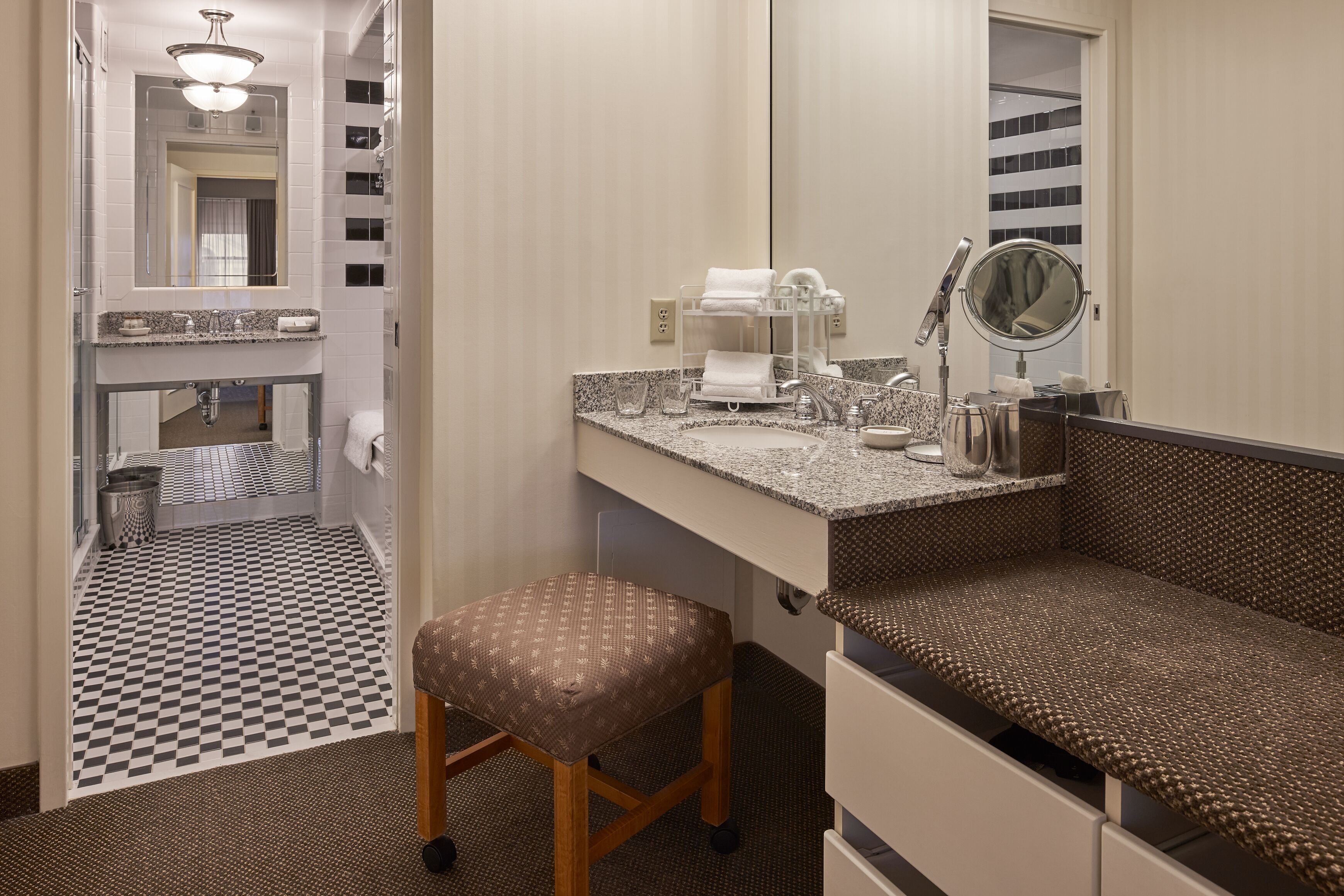 Une salle de bain d’hôtel avec un carrelage à motifs géométriques, un lavabo avec un miroir, un porte-serviettes et un banc.