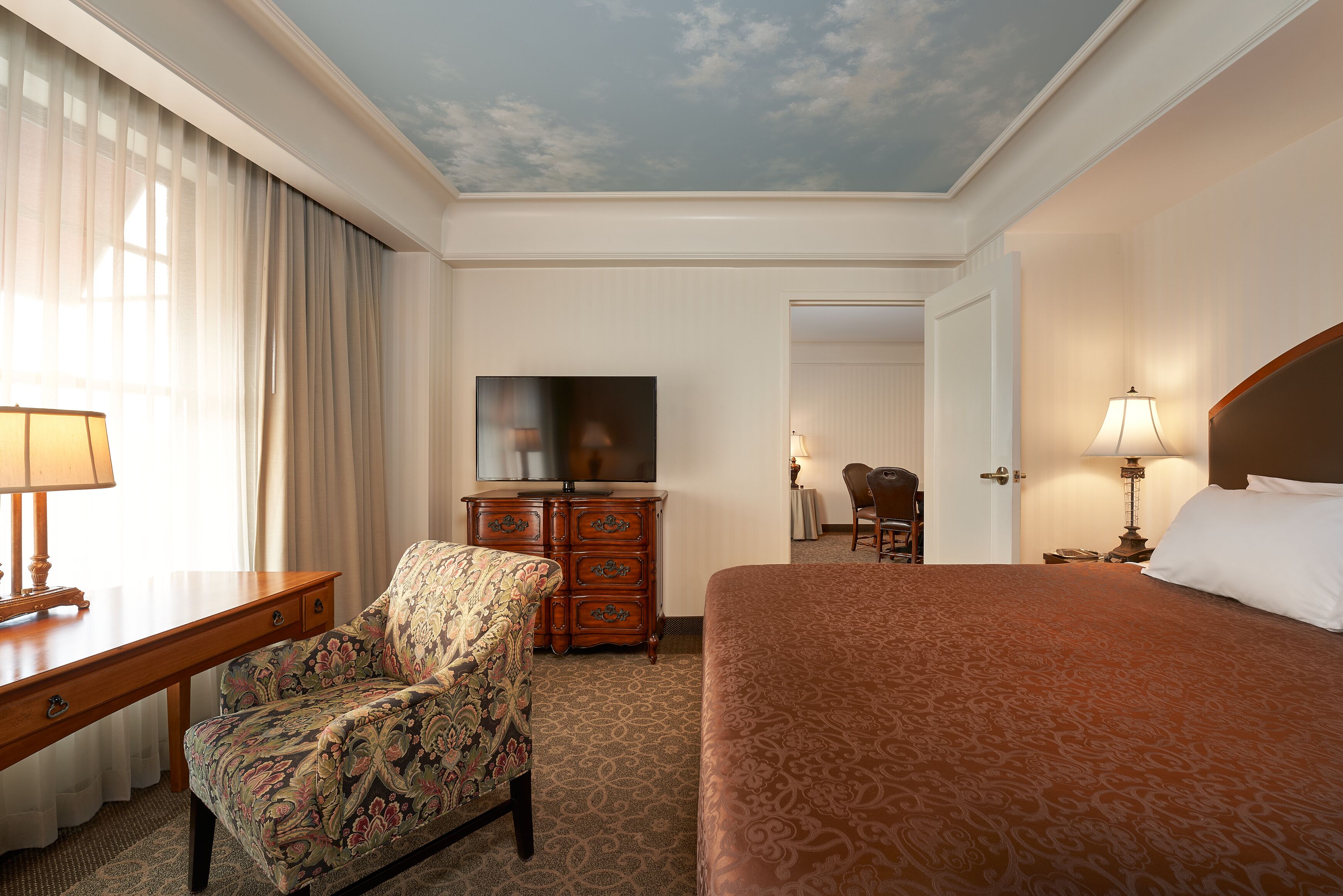 A hotel room with a bed, a desk with a lamp, a chair, a dresser, a television, and a door leading to another room.