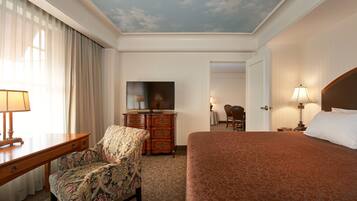 A hotel room with a bed, a desk with a lamp, a chair, a dresser, a television, and a door leading to another room.