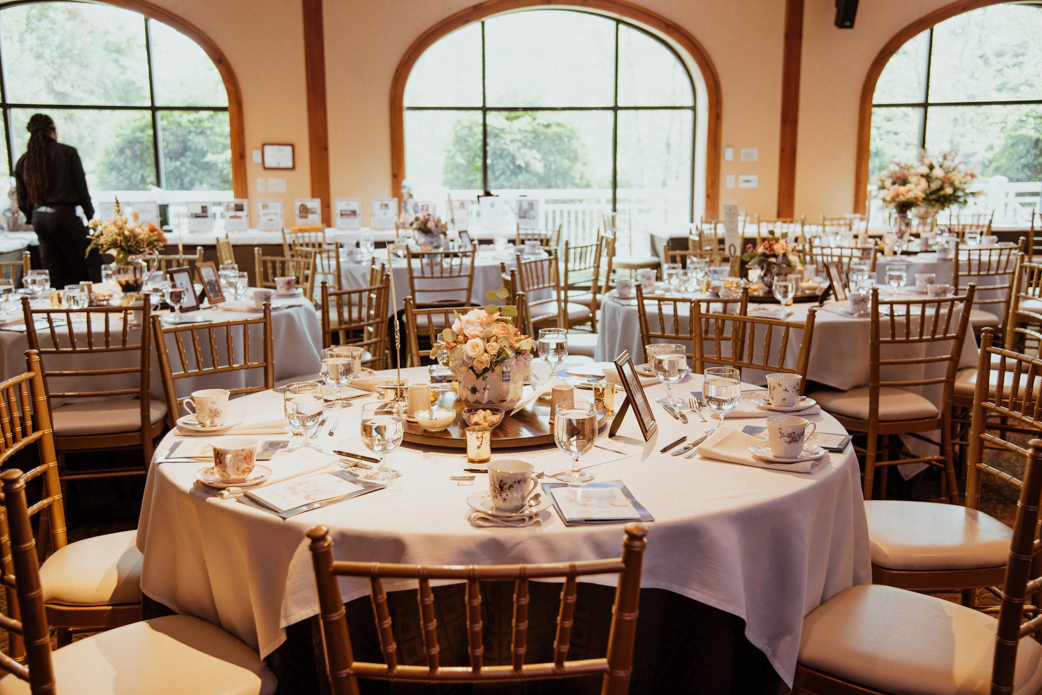 Un salón de banquetes con mesas redondas dispuestas para un evento formal, con manteles blancos, centros de mesa florales y sillas de madera.