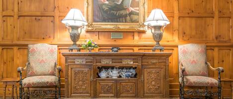 Une pièce avec un mur lambrissé en bois, un tableau encadré, deux chaises, un buffet contenant des services à thé et deux lampes.