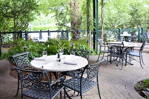 An outdoor patio with round tables and metal chairs, surrounded by greenery and a brick pathway.