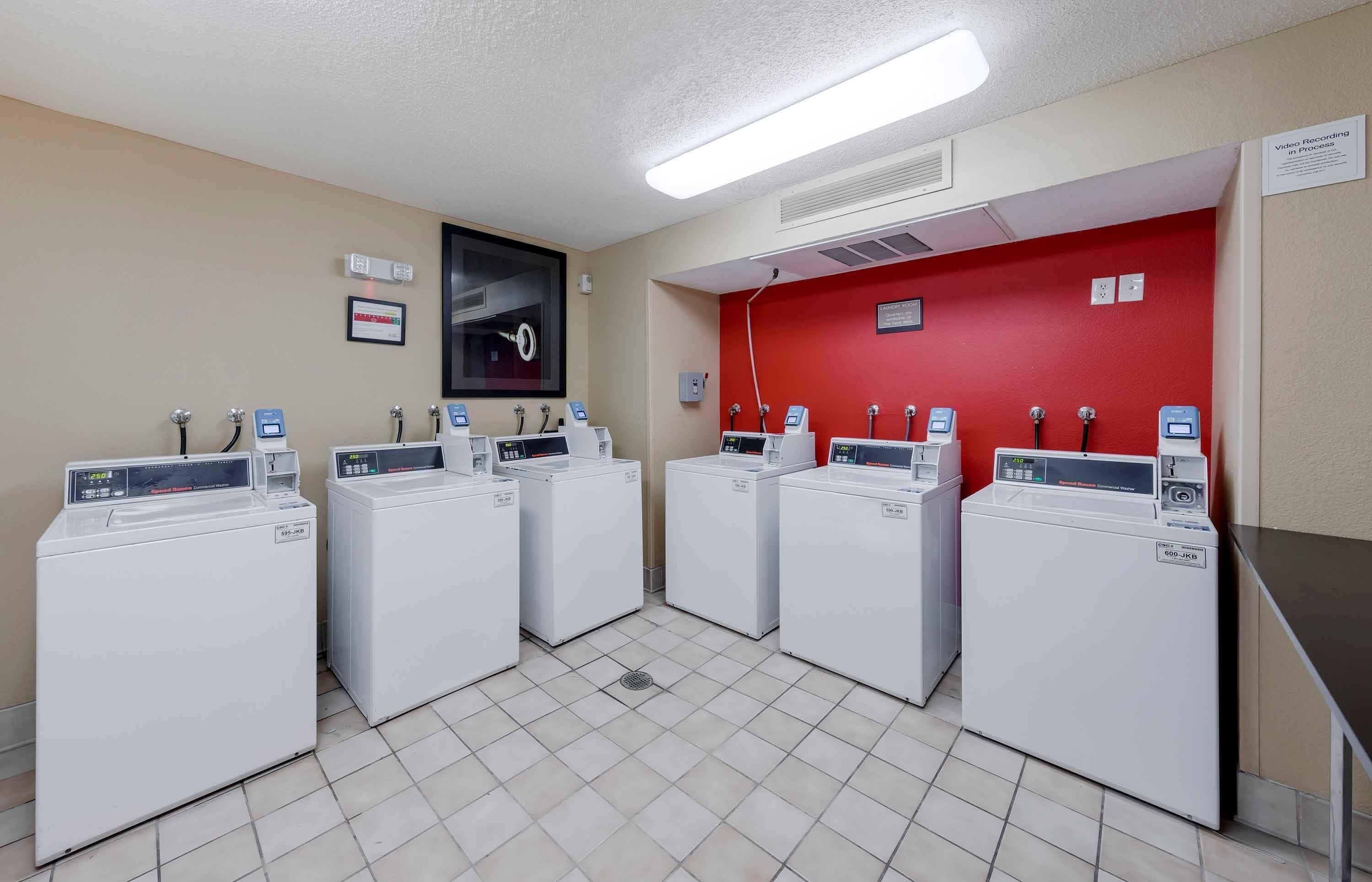 A laundry facility with four commercial washers and dryers, a red wall, and a sign indicating video recording in process.