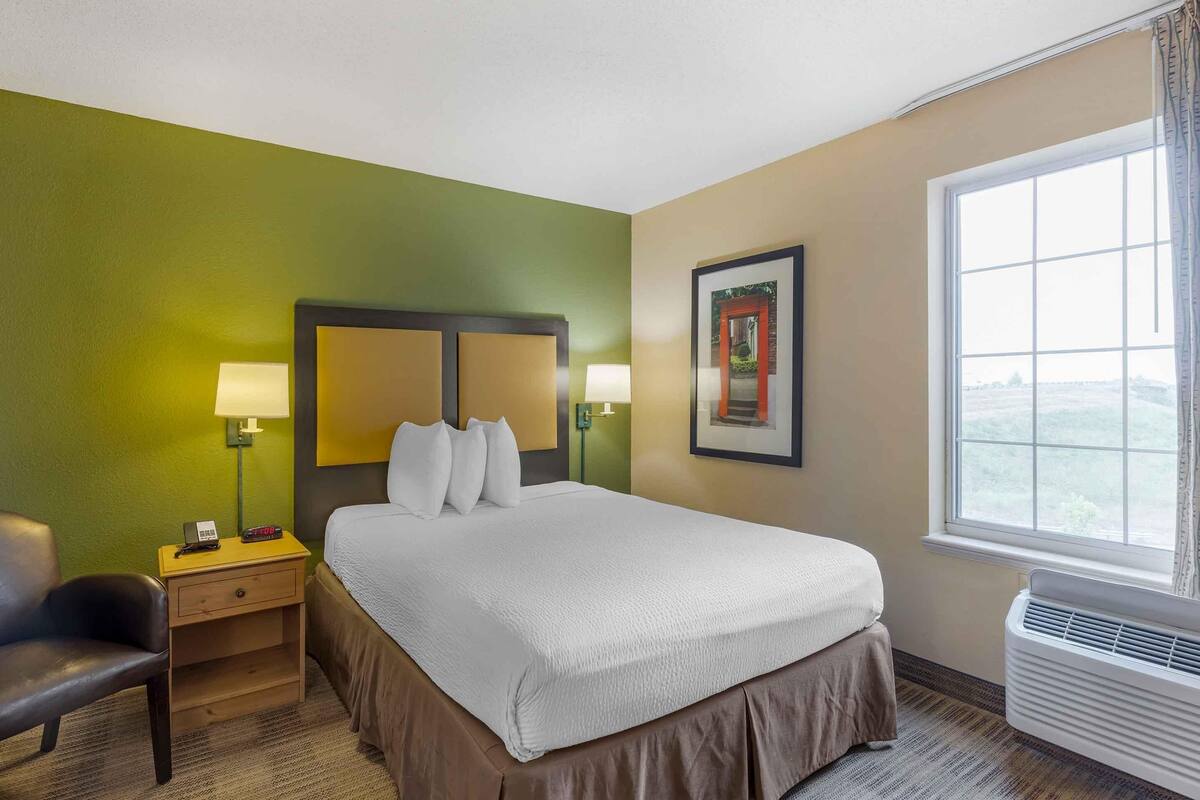 A hotel room with a bed, bedside lamps, a nightstand, a chair, a framed picture, and a window with a view.