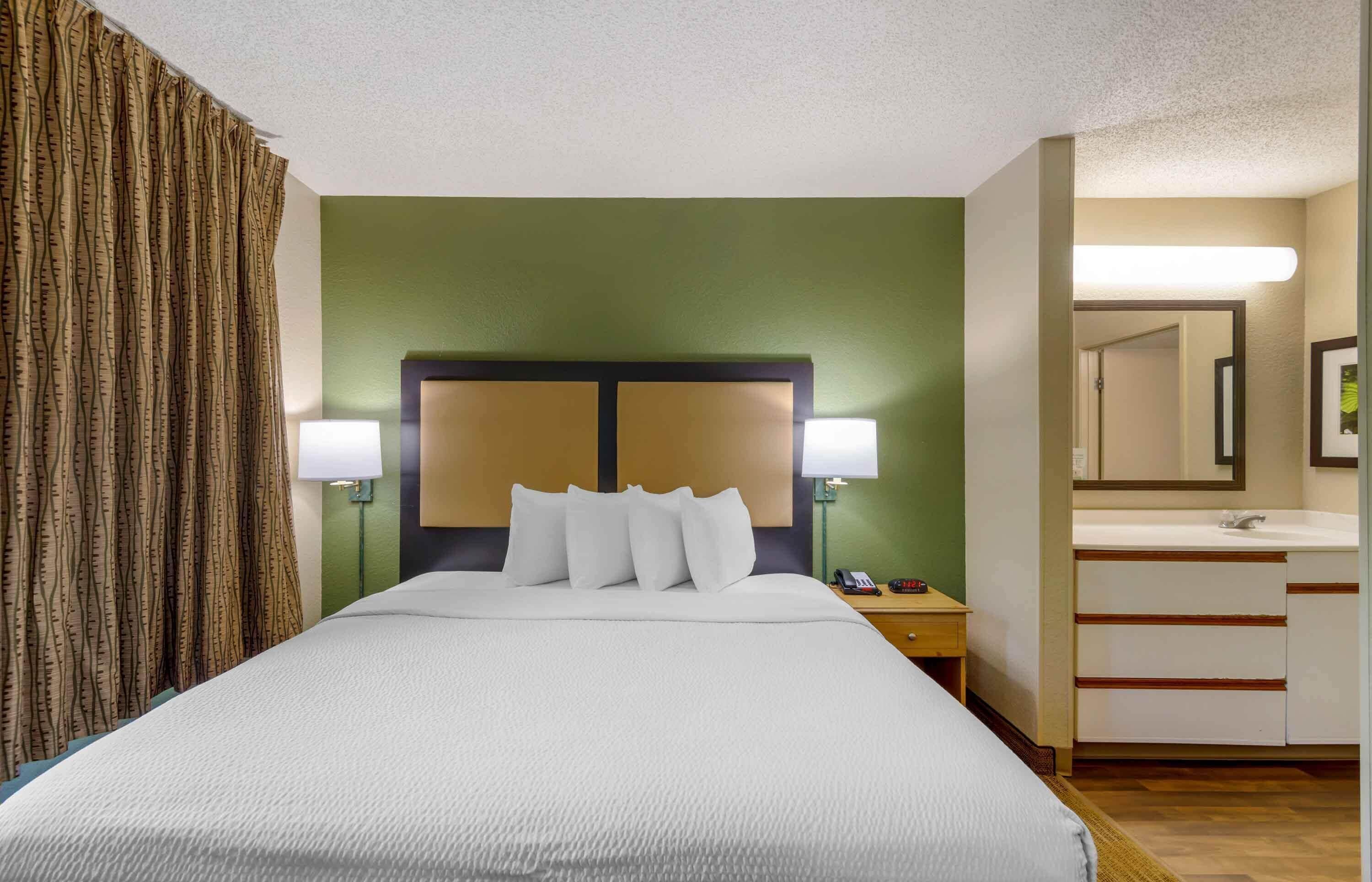 A hotel room with a large bed, two bedside lamps, a nightstand, a mirror, and a bathroom with a sink and mirror.
