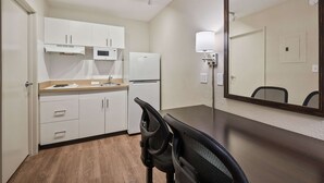 Una cocina compacta con armarios blancos, microondas y refrigerador. Hay una mesa de comedor con sillas y un espejo grande en la pared.