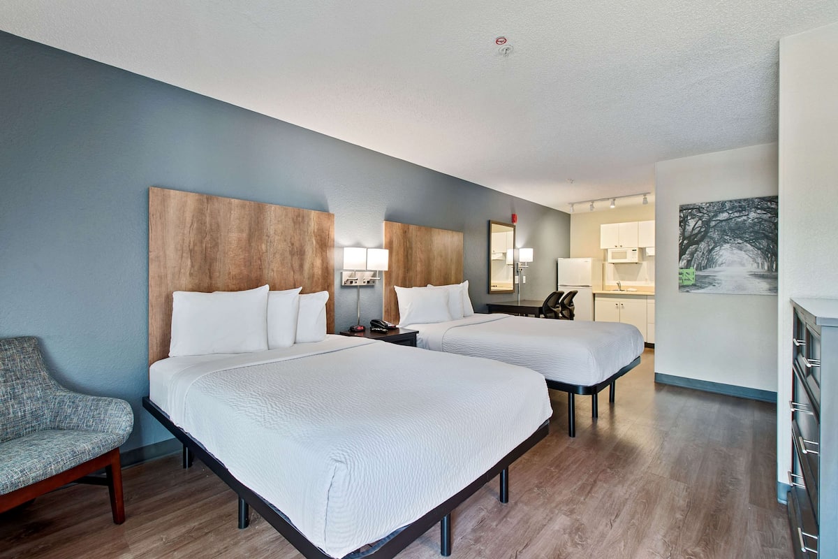 A hotel room with two beds, a chair, a desk, and a kitchenette.