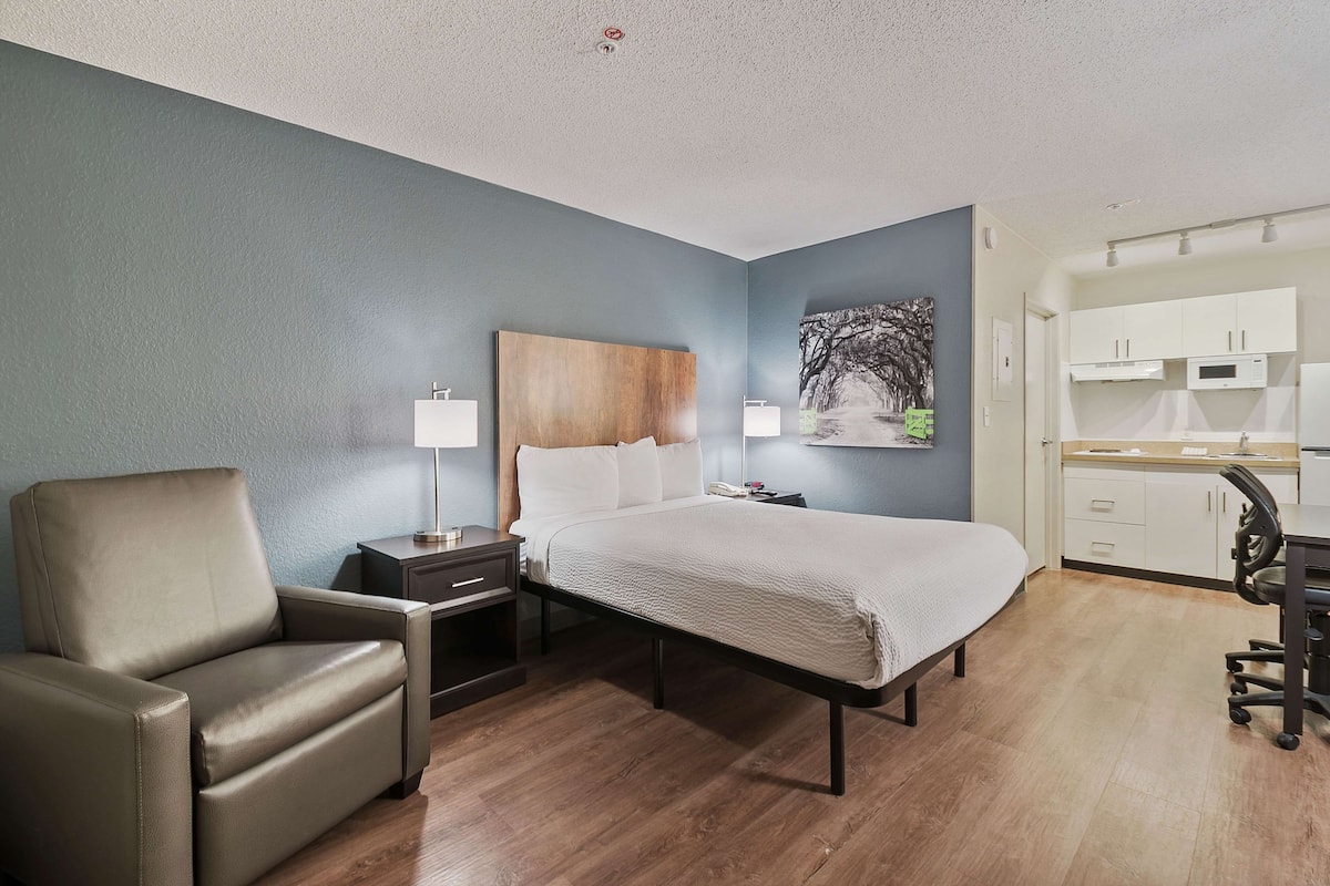 A hotel room with a bed, a chair, a desk, a nightstand, a lamp, a painting, and a kitchenette.