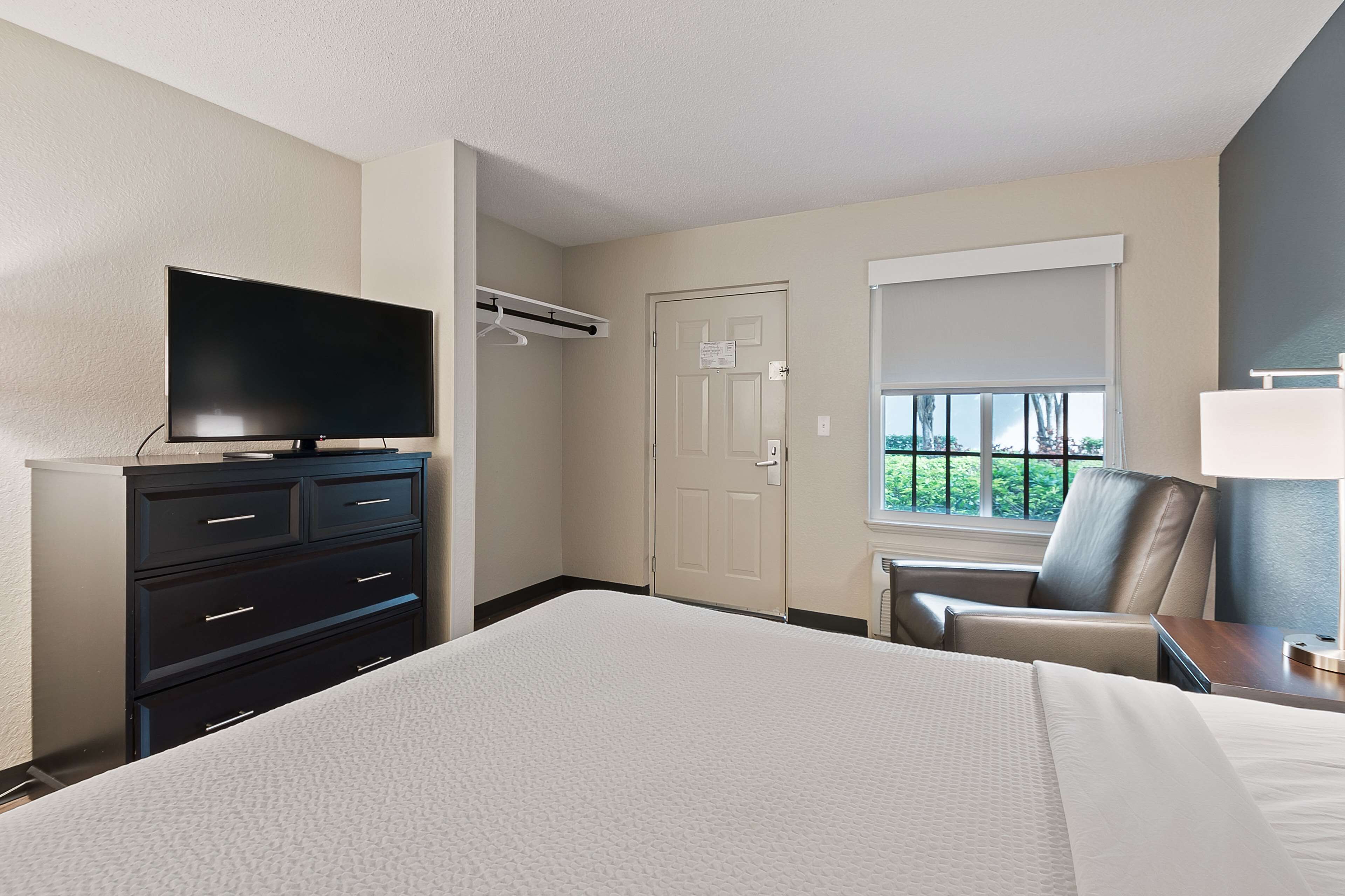 A hotel room with a large bed, a flat-screen TV on a dresser, a chair, a desk with a lamp, and a window with a view of greenery.