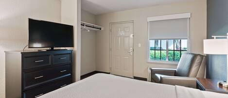 A hotel room with a large bed, a flat-screen TV on a dresser, a chair, a desk with a lamp, and a window with a view of greenery.