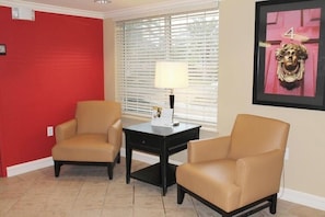 A room with two beige armchairs, a small black table with a lamp, a window with blinds, and a framed picture on the wall. - Studio 6 Extended Stay Memphis Airport TN (Memphis)