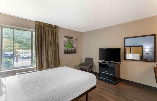 A hotel room with a bed, a television, a chair, a dresser, a window with curtains, and a painting on the wall. - Studio 6 Extended Stay Memphis Airport TN (Memphis)