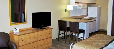 A compact hotel room with a single bed, a television on a wooden dresser, a small kitchenette with a refrigerator, and a mirror above the dresser.