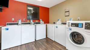 A laundry room with multiple washing machines and dryers, a television mounted on the wall, and a window with blinds. - Extended Stay America Select Suites - Washington, DC - Sterling - Dulles (Sterling)