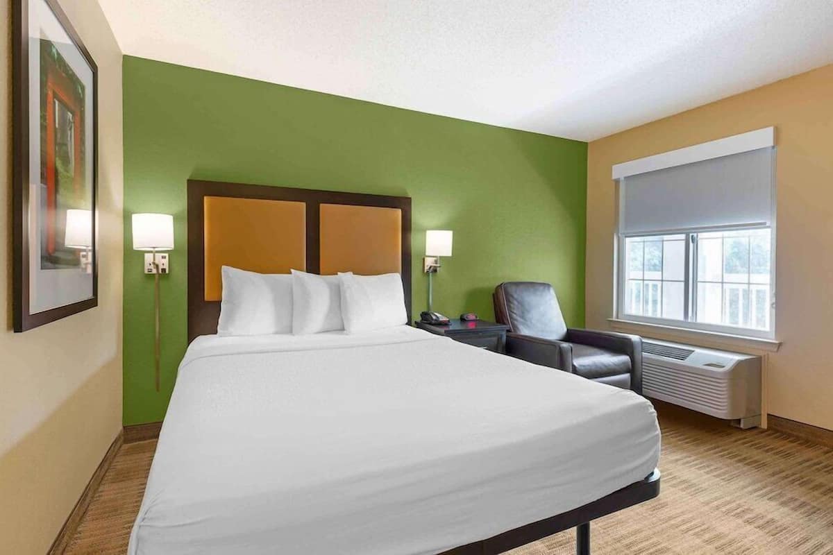 Une chambre d’hôtel avec un grand lit, des murs verts, une fenêtre avec des stores, une chaise et un radiateur.