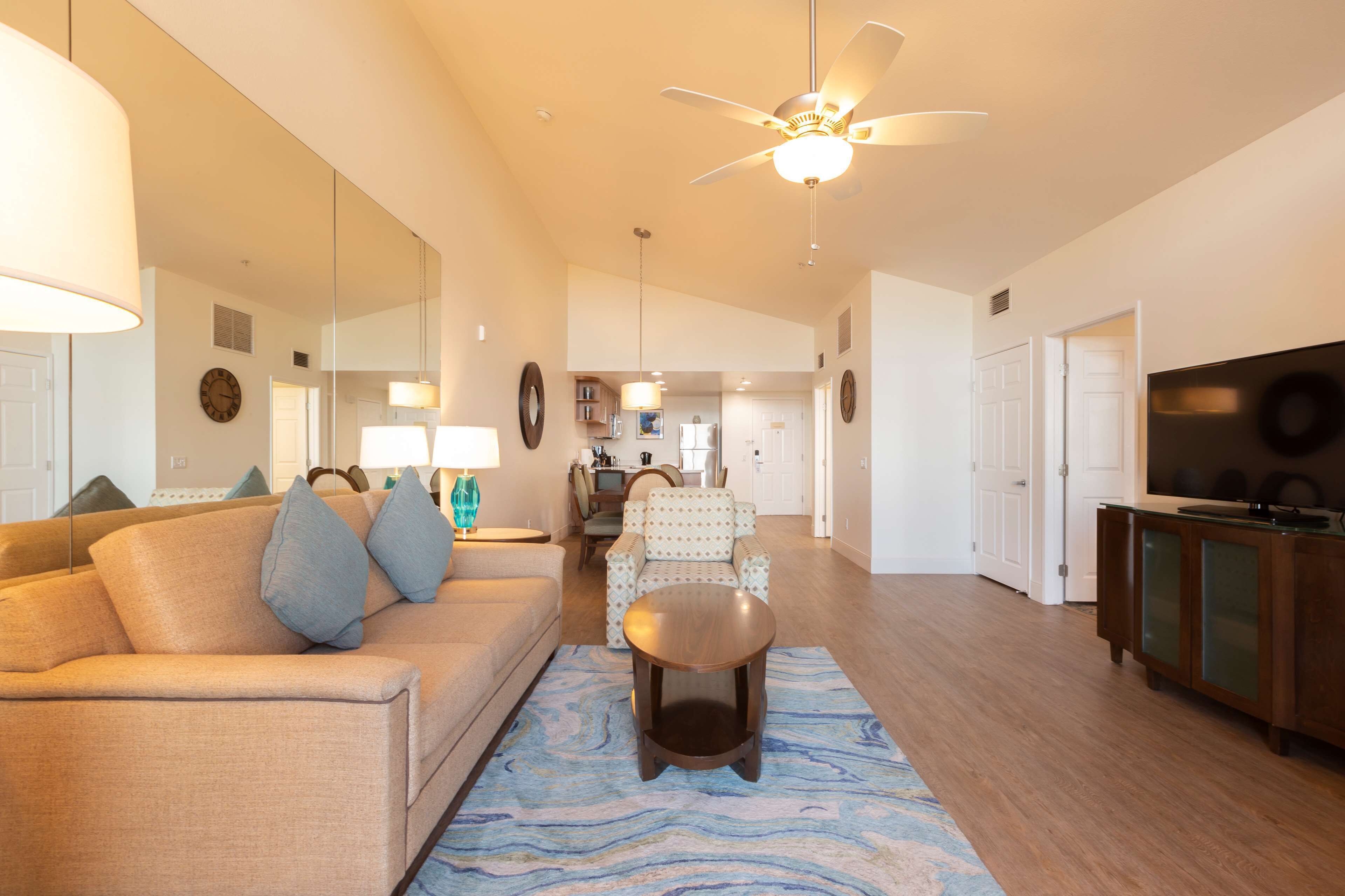 A spacious living room with a sofa, a coffee table, a ceiling fan, and a kitchen in the background.