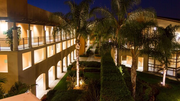 A hotel courtyard with a covered walkway, palm trees, and well-lit building facades. - Grand Pacific Palisades (Carlsbad)