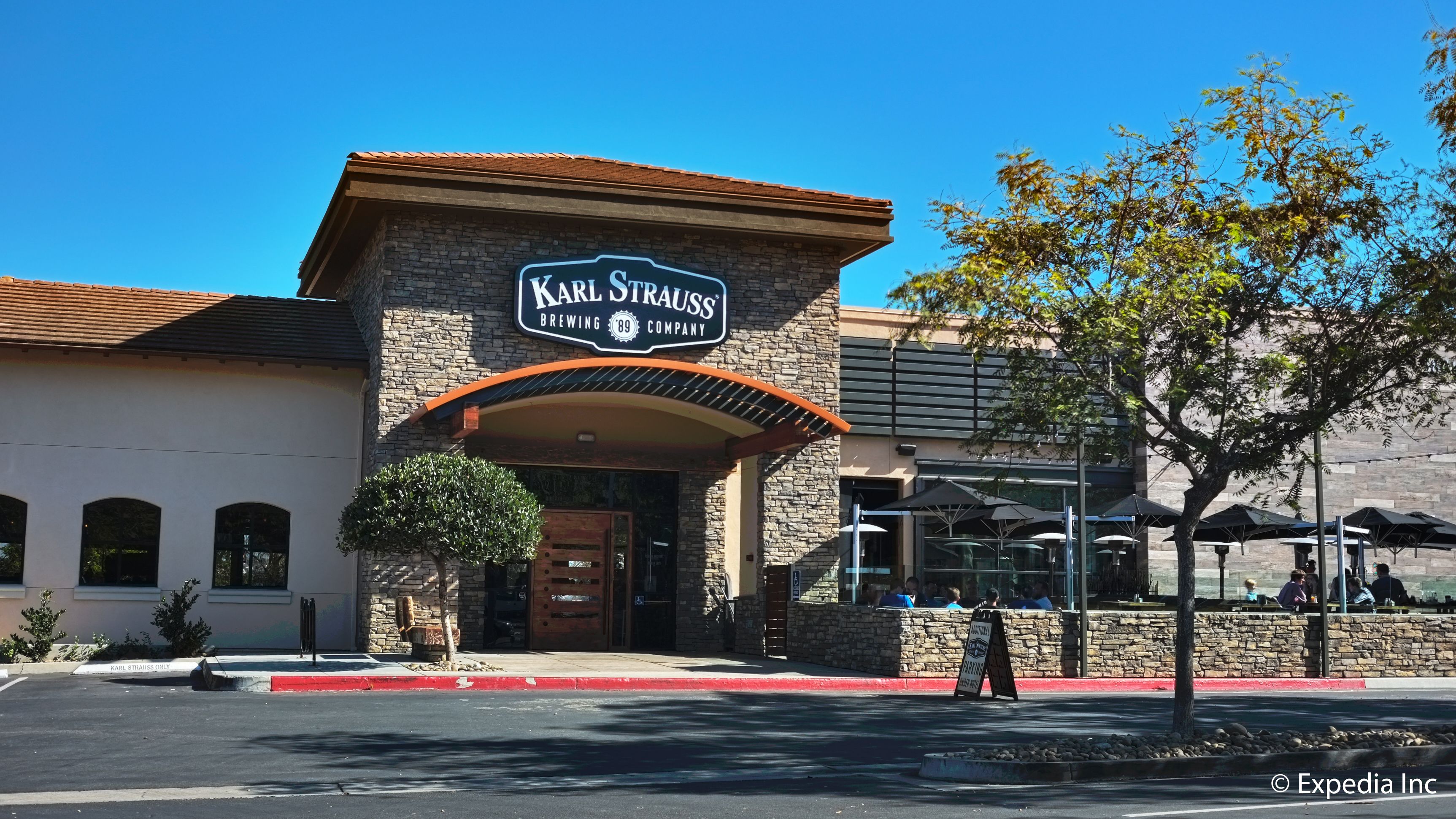 A Karl Strauss Brewing Company building with a stone facade, a red awning, and outdoor seating with umbrellas.