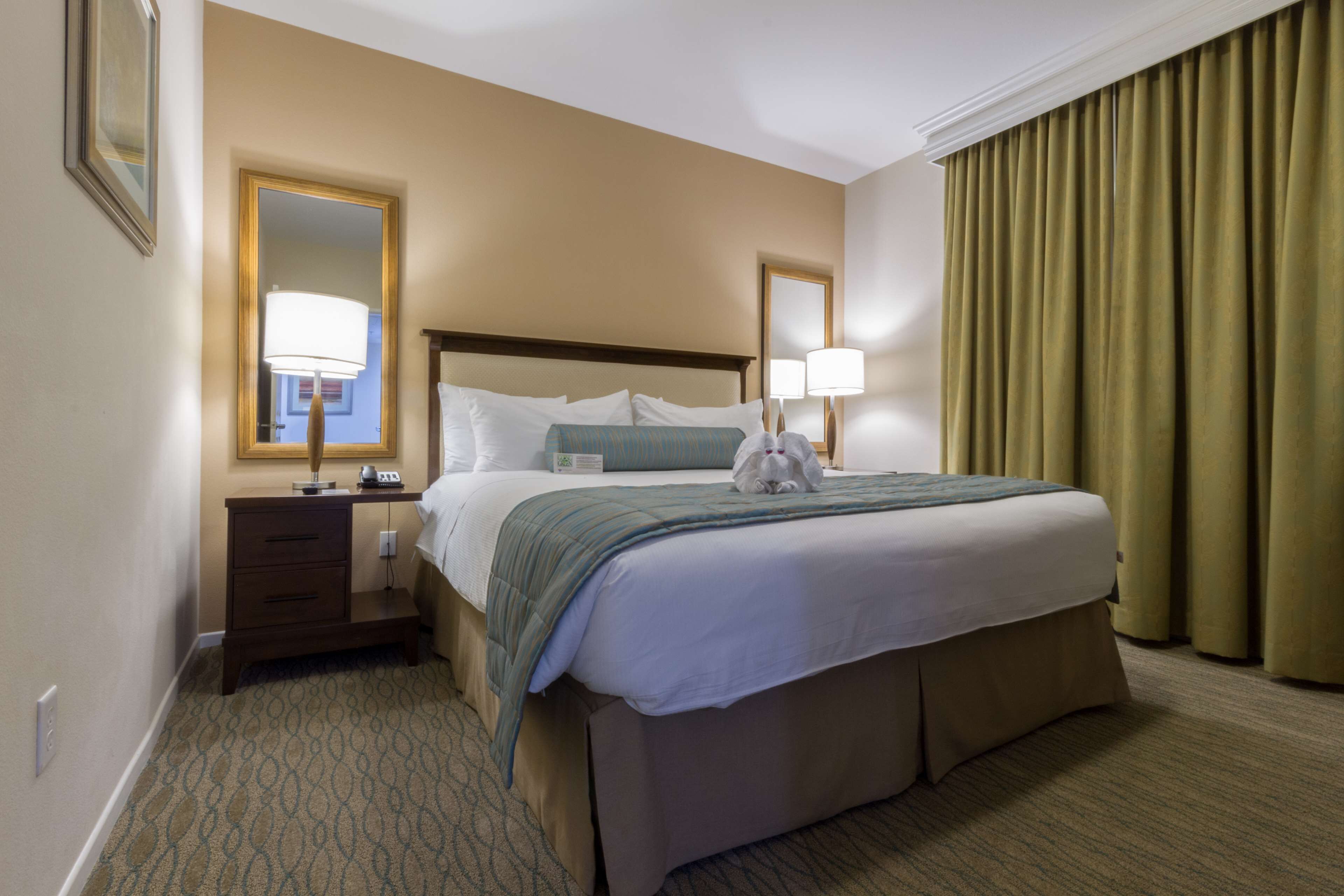 Quarto de hotel com uma cama grande, duas luminárias de cabeceira, mesa de cabeceira, espelho e janela com cortinas.