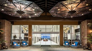 A lobby with a modern ceiling design, glass doors leading to a seating area with cushioned chairs, and a brick floor. - Grand Pacific Palisades (Carlsbad)
