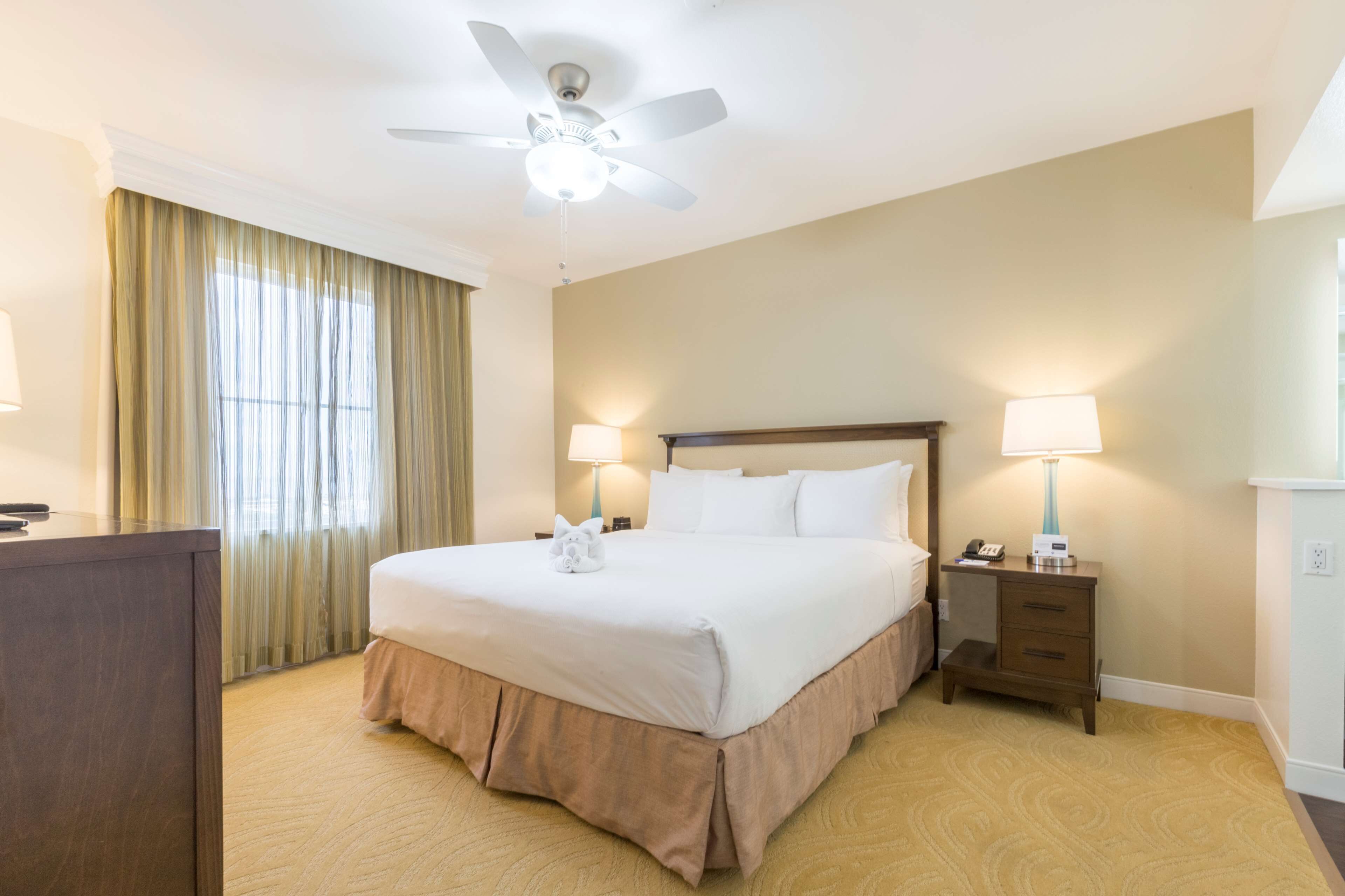 Habitación de hotel con una cama grande, dos lámparas de noche, una mesita de noche, un escritorio, un ventilador de techo y una ventana con cortinas.