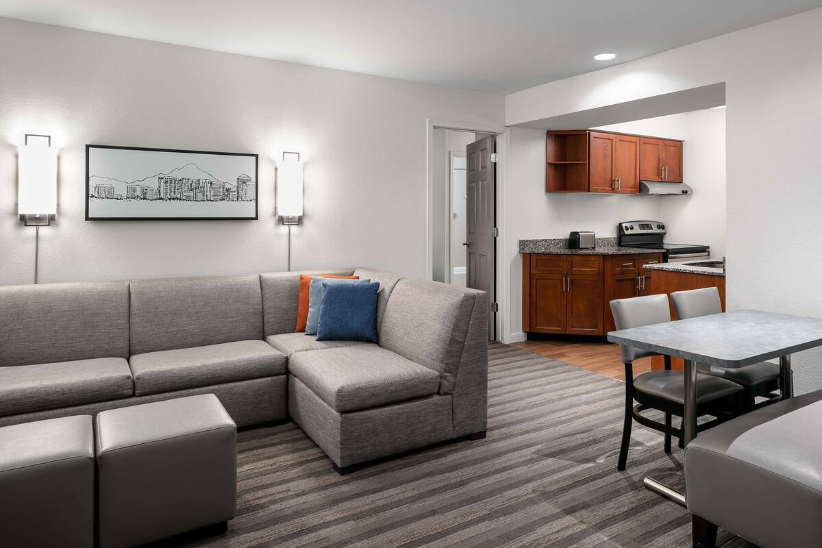 A modern living room with a sectional sofa, a dining table, and a kitchen area with wooden cabinets and appliances.