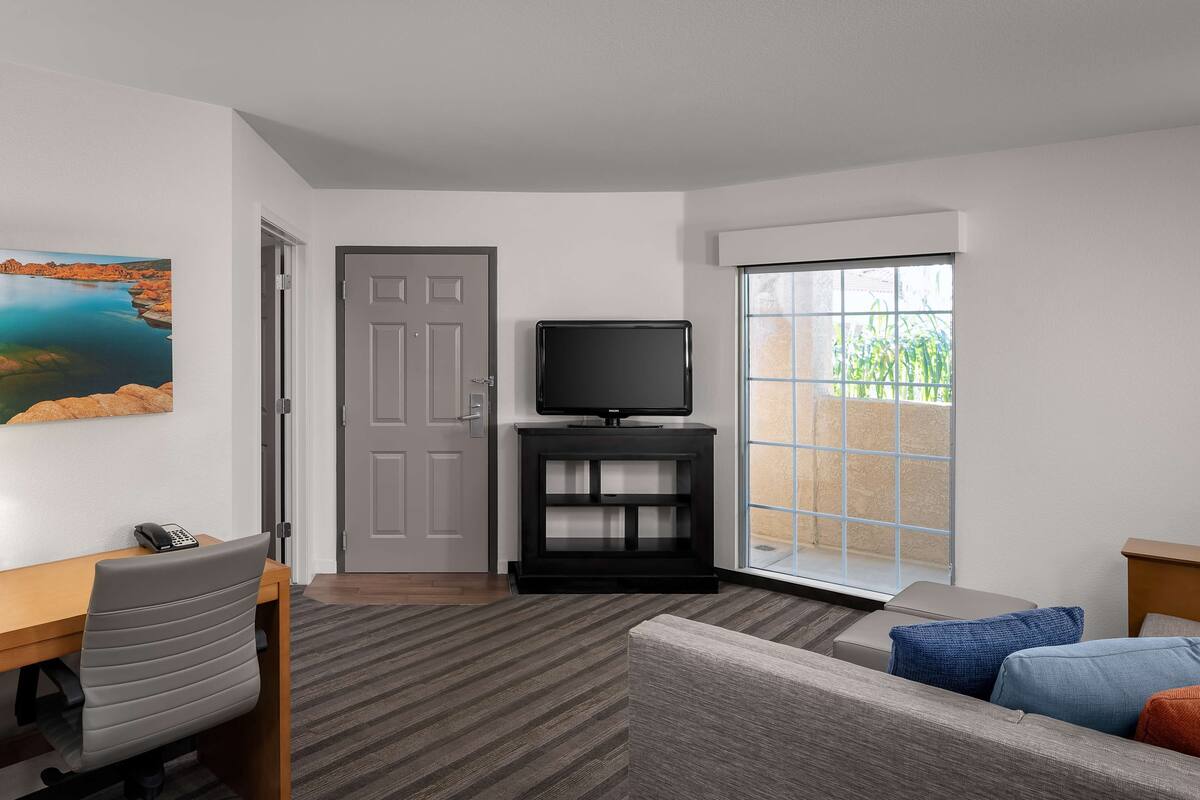 A hotel room with a desk, a television on a black stand, a sofa with cushions, a large window with a view of greenery, and a door.