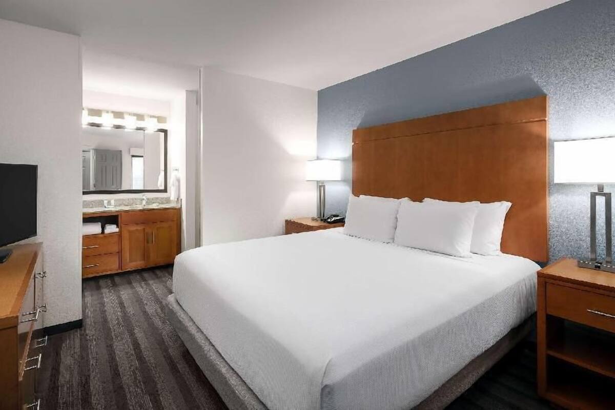 A hotel room with a large bed, wooden headboard, bedside tables, a TV, a bathroom with a mirror, and a wooden floor.
