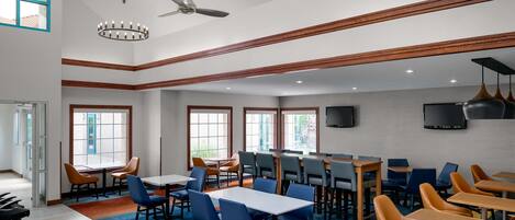 A spacious dining area with multiple tables and chairs, large windows, ceiling fans, and a flat-screen TV mounted on the wall.