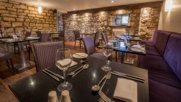 Un restaurante con paredes de piedra, suelos de madera y asientos tapizados en color morado. Las mesas están dispuestas con copas de vino, platos y cubiertos.