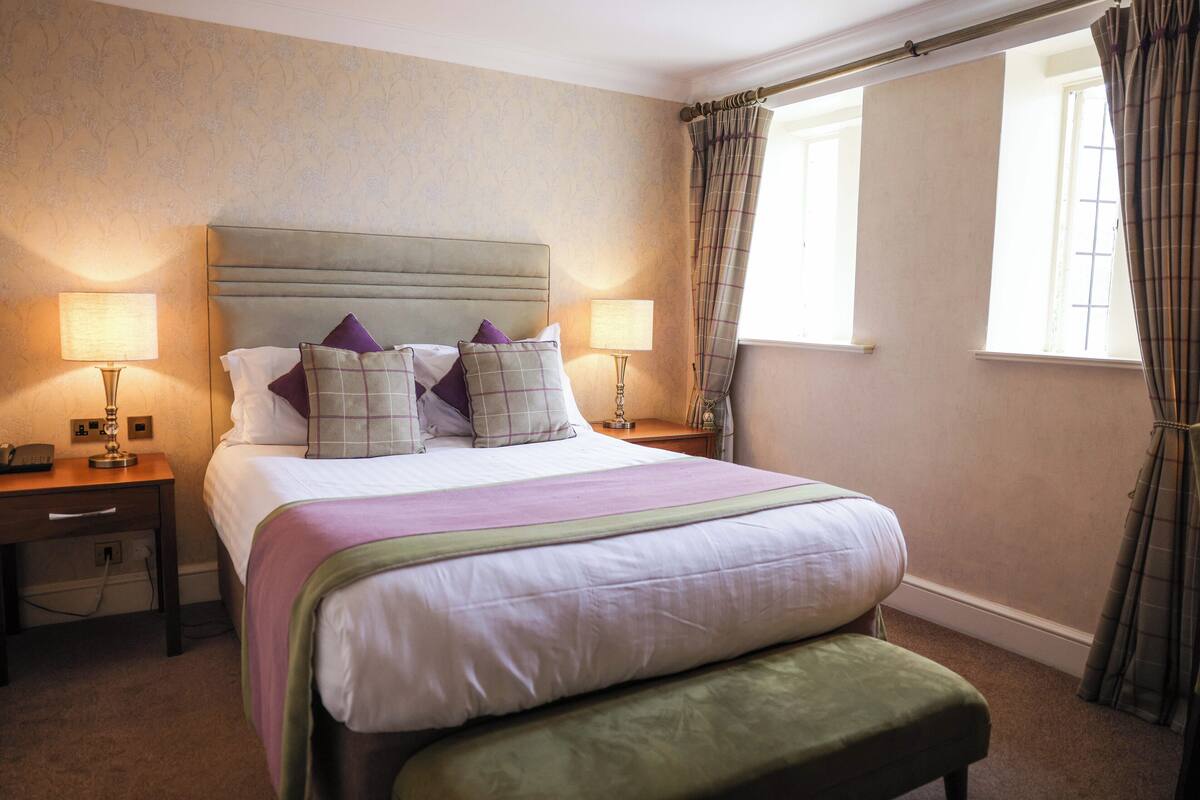 A neatly made bed with a headboard, two bedside tables with lamps, a bench, and a window with curtains.