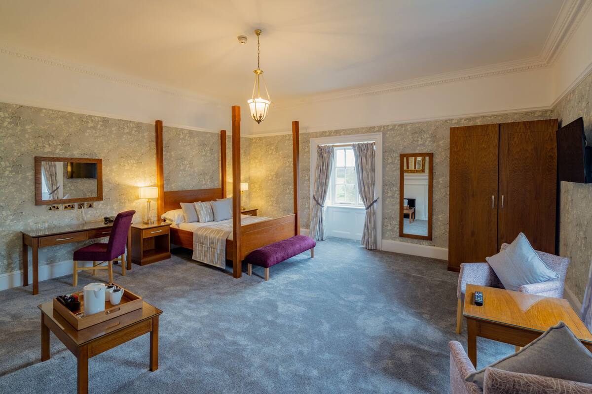 A spacious bedroom with a four-poster bed, a desk, a chair, a sofa, and a TV.
