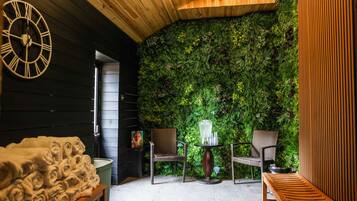Une pièce chaleureuse avec un plafond en bois, un mur recouvert de mousse verte, deux fauteuils en osier, un banc en bois et une horloge.