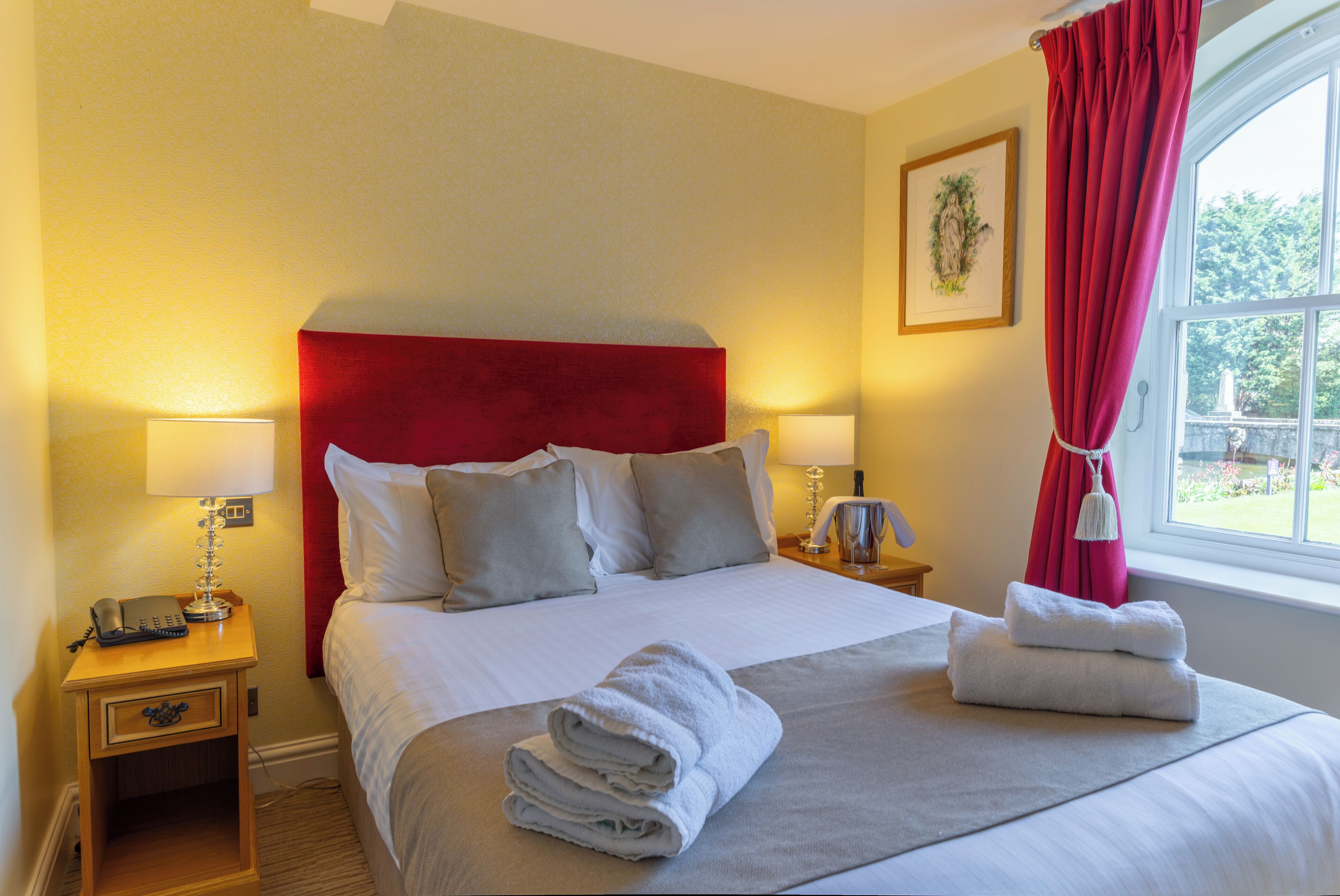 A hotel room with a large bed, two bedside tables, a red headboard, and a window with a view of greenery.