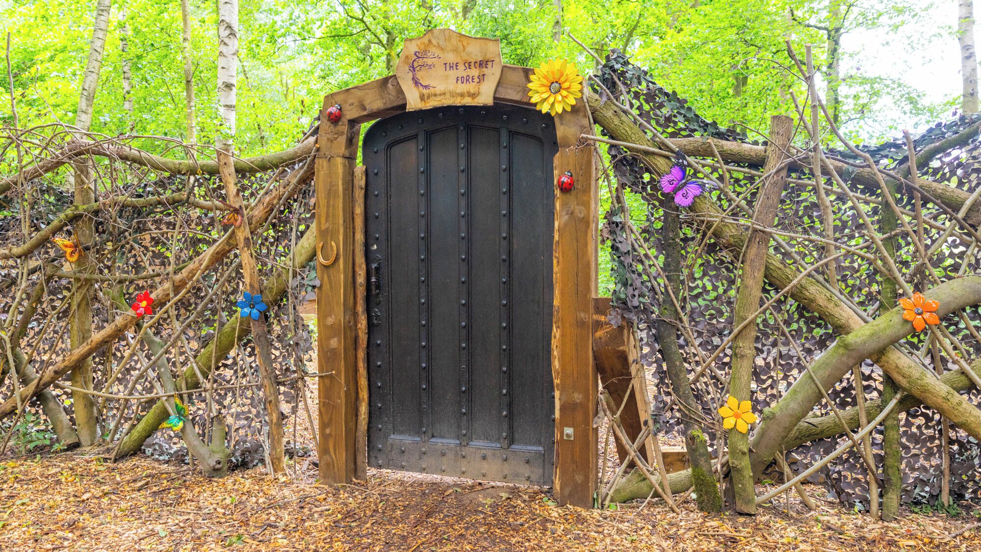 Una reja de madera con un letrero que dice 'The Secret Forest' en un entorno natural con árboles y follaje.