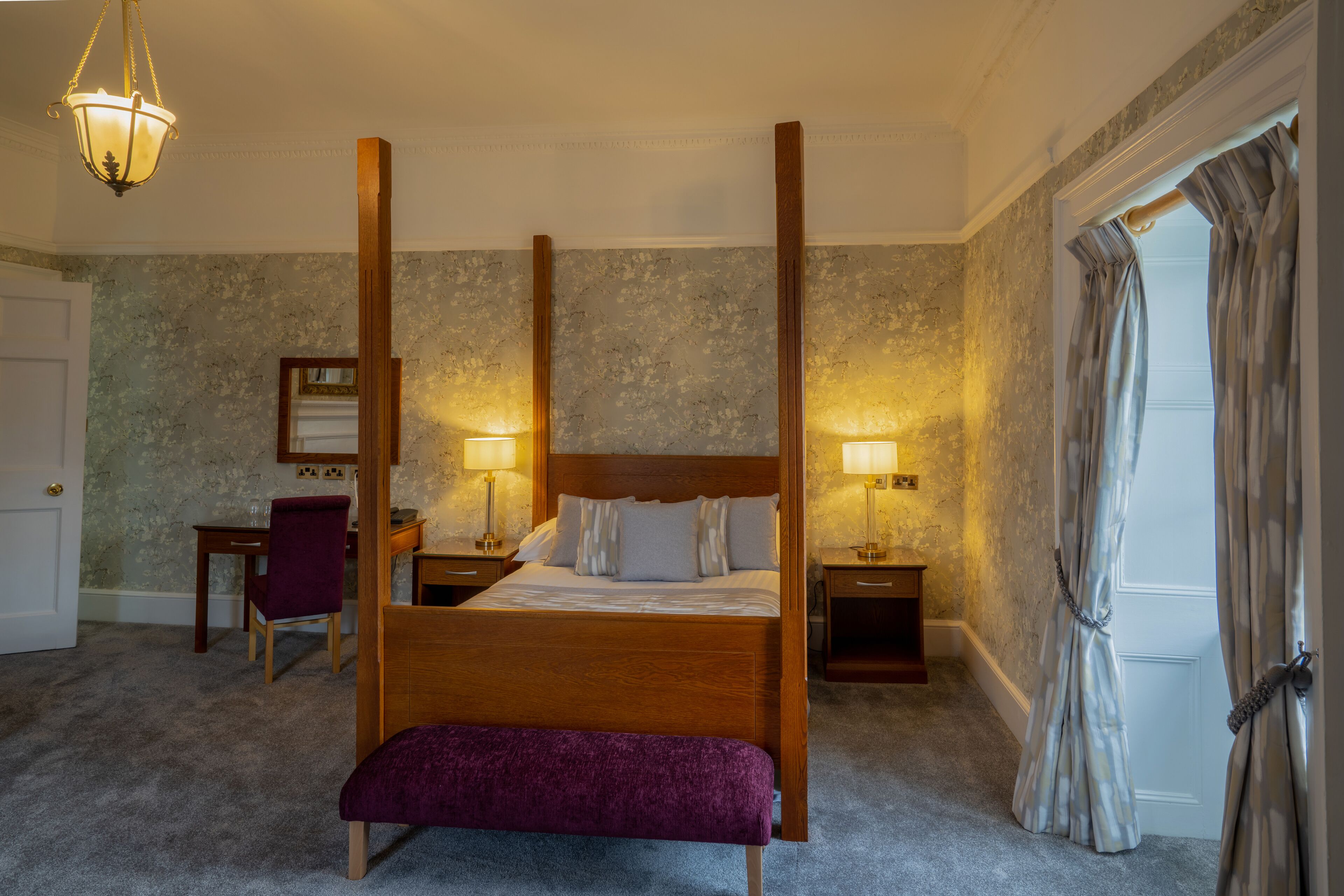 A four-poster bed with a wooden headboard, two bedside tables with lamps, a purple bench, a desk with a chair, and a large mirror on the wall.