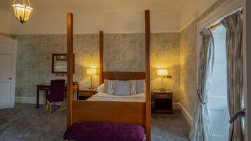 Ein Bett mit Baldachin, ein Holzkopfstück, zwei Nachttische mit Lampen, eine violette Bank, ein Schreibtisch mit Stuhl und ein großer Spiegel an der Wand.