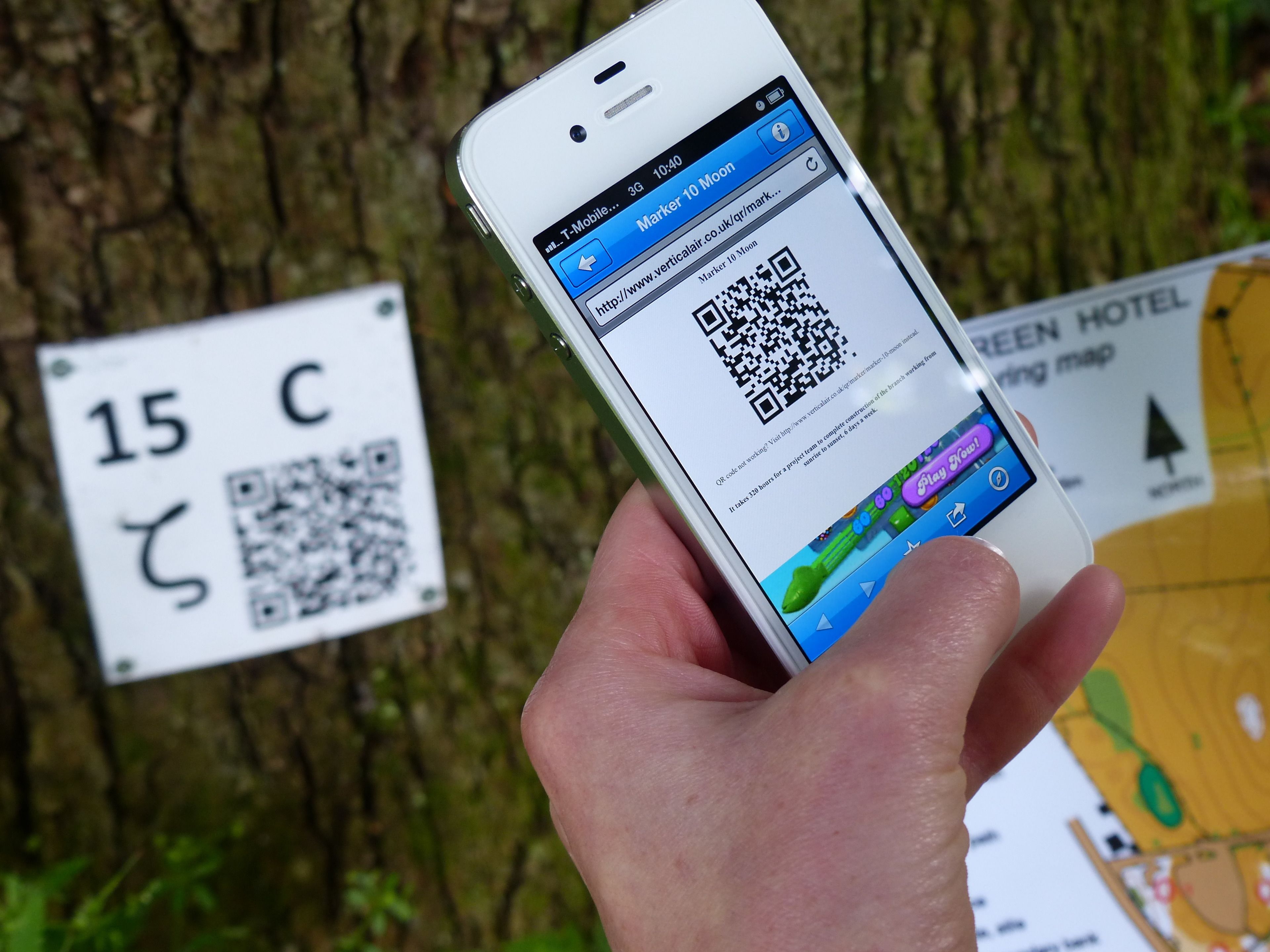 Une main tenant un smartphone affichant un code QR et l’adresse d’un site web. En arrière-plan, on aperçoit un panneau avec un code QR et une carte.