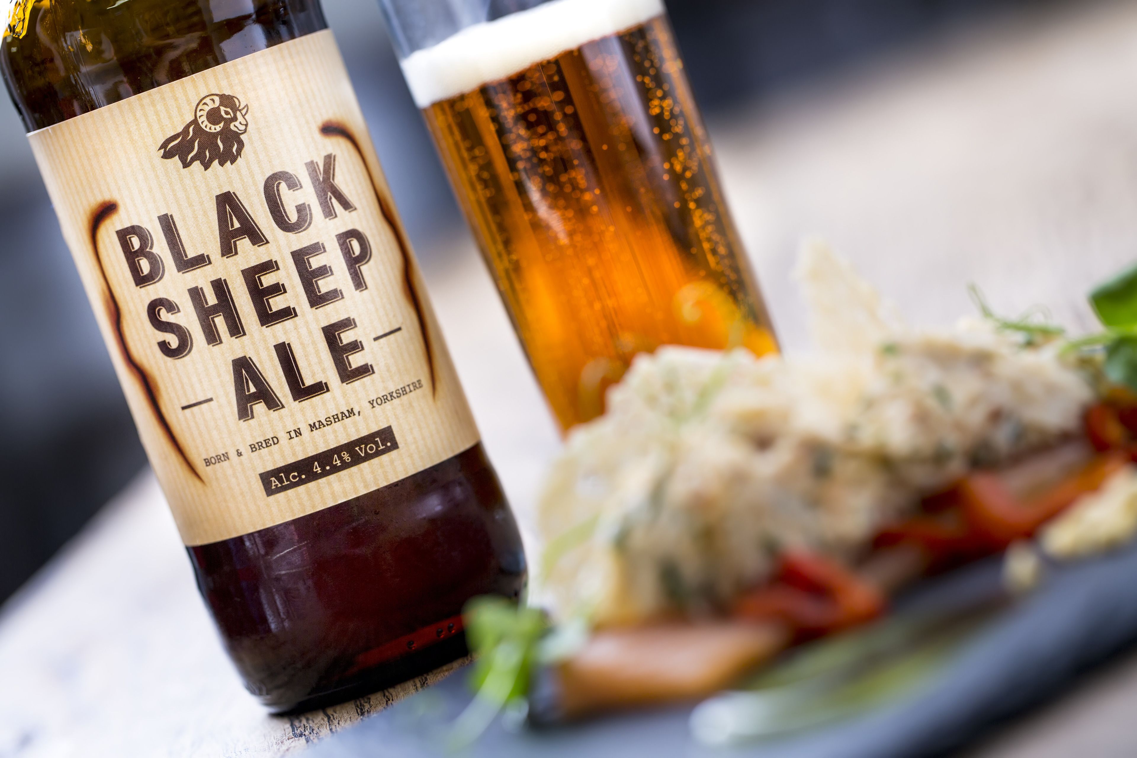Une bouteille de bière Black Sheep, avec son étiquette, et un verre de bière, placés à côté d’une assiette de nourriture.