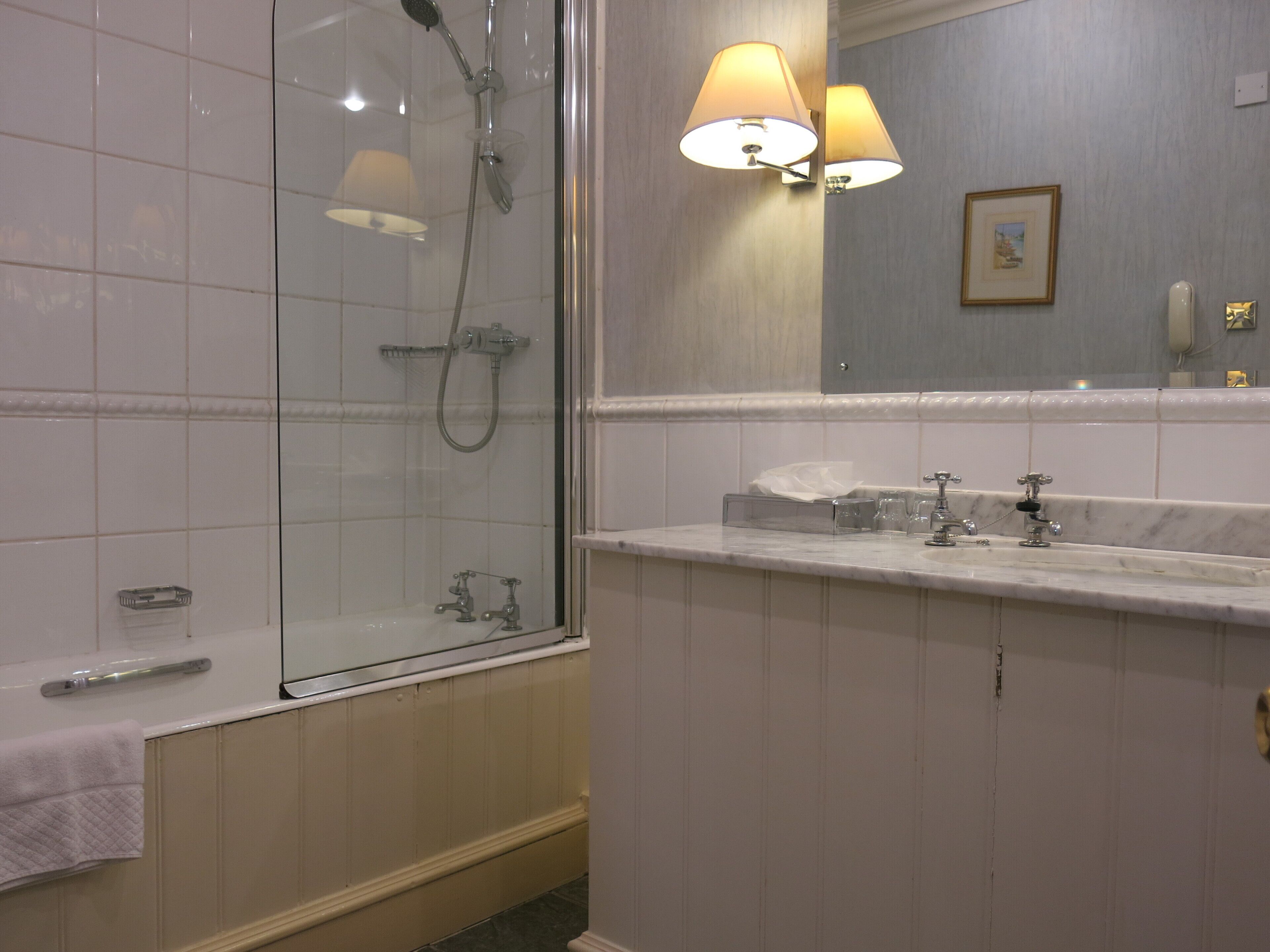 Une salle de bain dotée d’une douche à parois de verre, d’un meuble-lavabo avec un plan de travail en marbre et de deux lampes fixées au mur.