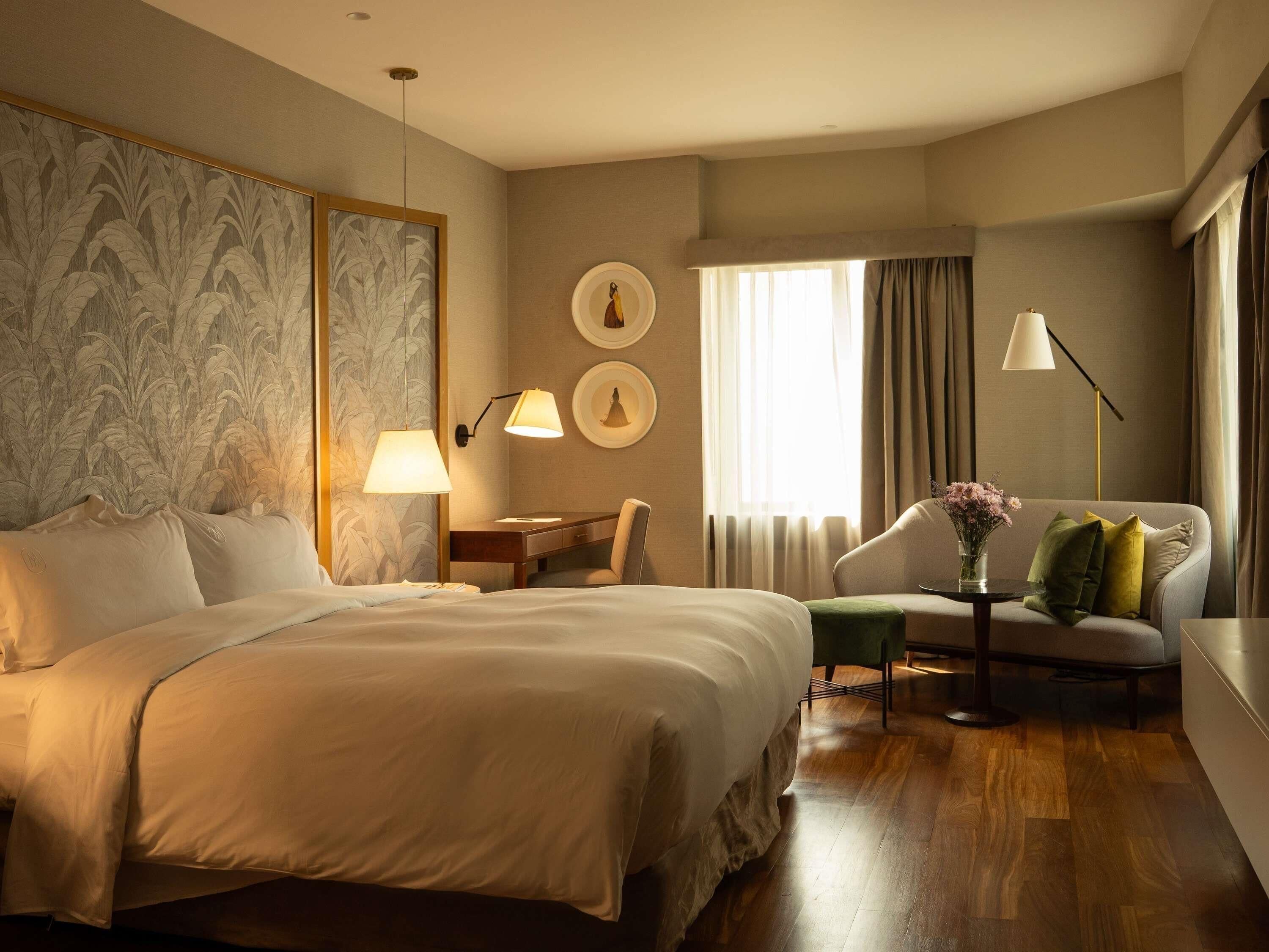 Habitación de hotel con una cama grande, un escritorio, una silla, una mesita con un florero y una ventana con cortinas.