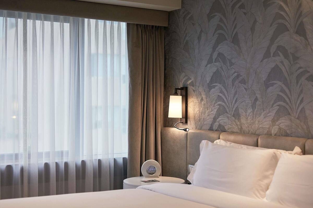 A hotel room with a bed, a nightstand, a lamp, and a window with sheer curtains.