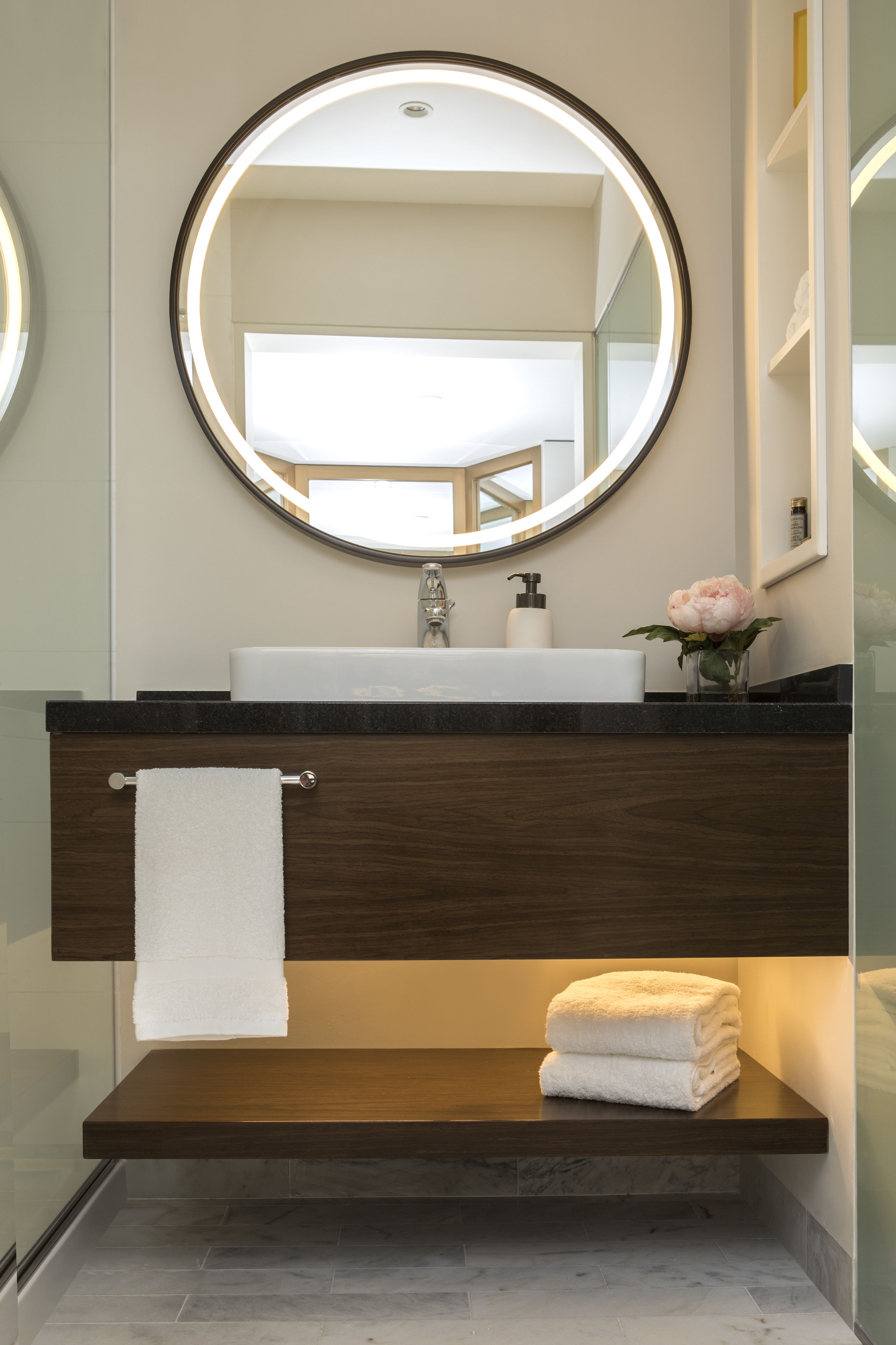 Une salle de bain moderne avec un miroir rond, un lavabo, un porte-serviettes et une étagère sur laquelle est posée une serviette pliée.
