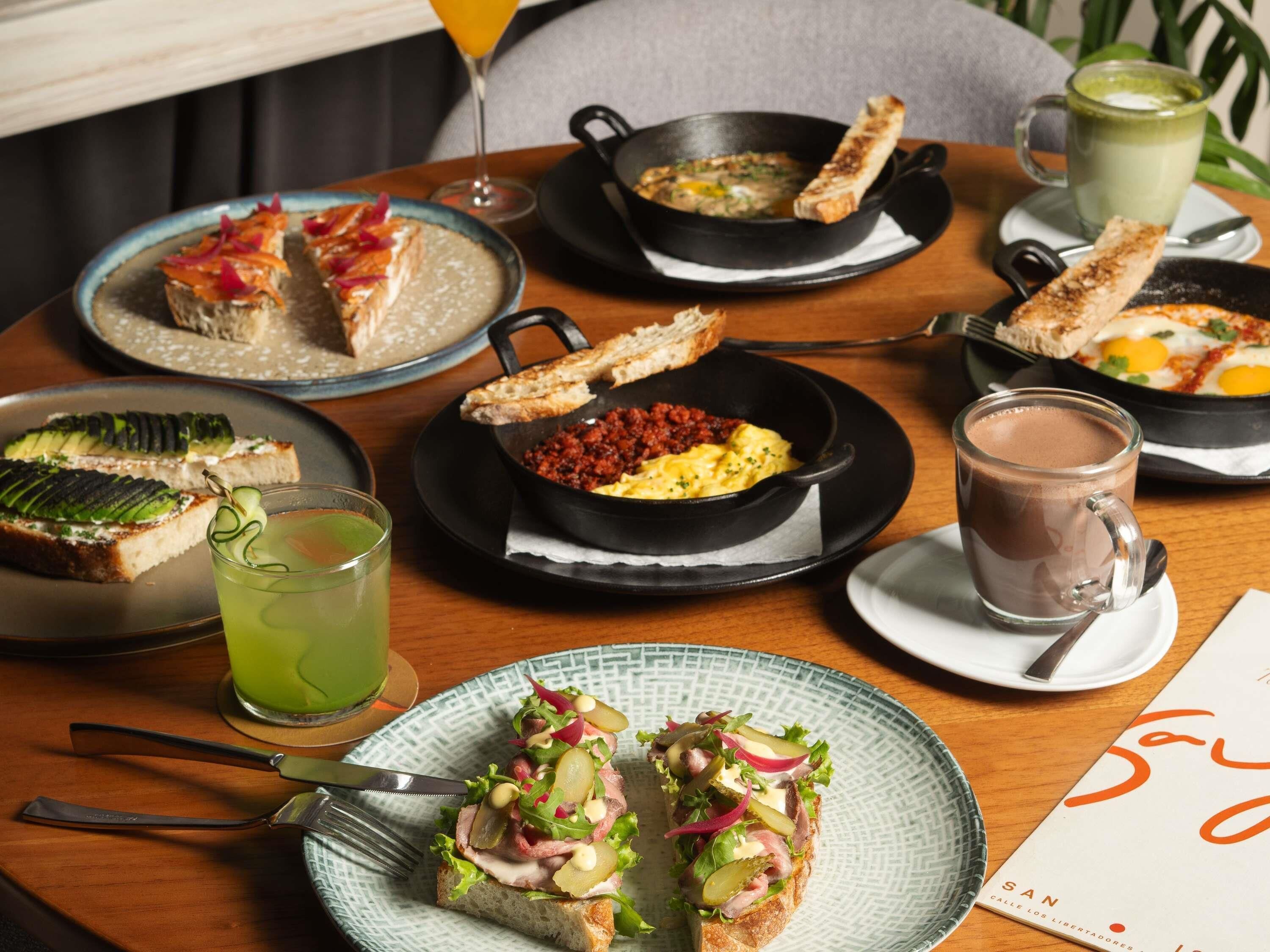 Una mesa de comedor con diversos alimentos para el desayuno, como tostadas, huevos revueltos y una bebida verde.