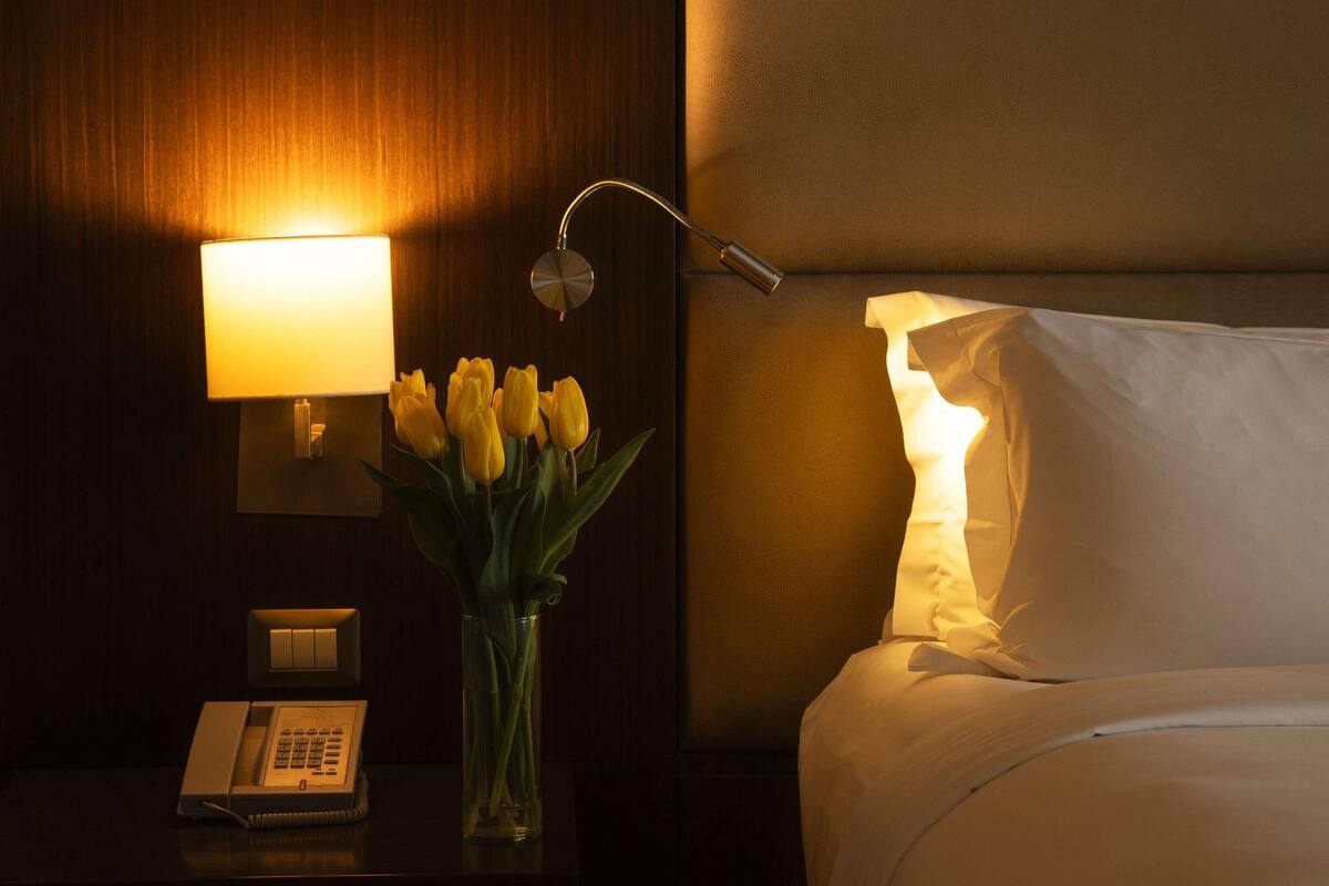 A hotel room with a bedside lamp, a telephone, a vase with flowers, and a bed with a pillow.